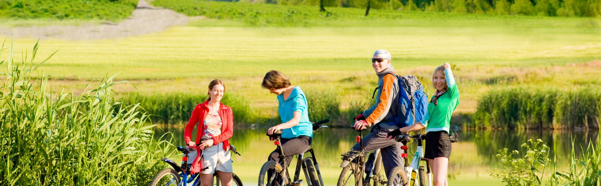 Eine vierköpfige Familie steht mit Fahrrädern an einem See, umgeben von grünen Wiesen und bewaldeten Bergen unter blauem Himmel. Sie machen eine Pause während ihrer Fahrradtour.