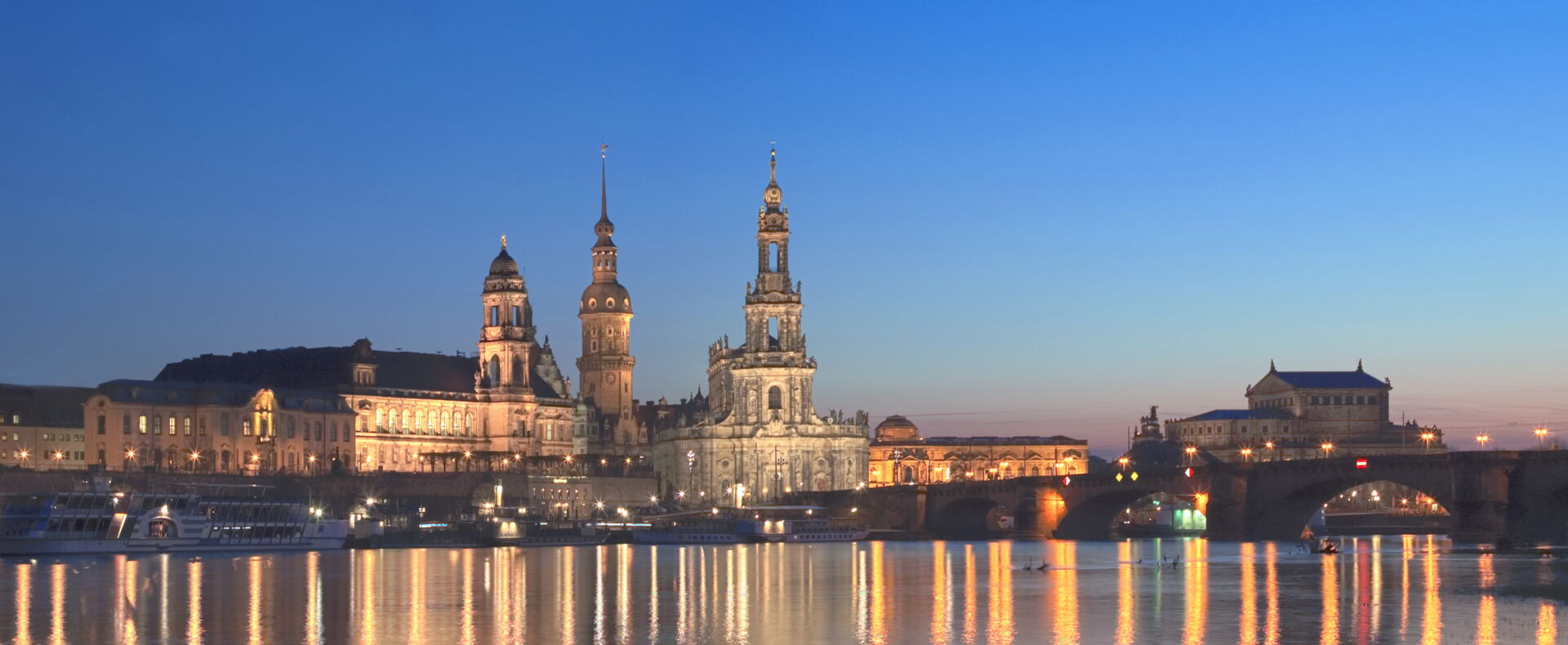Eine unscharfe Nachtaufnahme der Dresdner Skyline mit beleuchteten Gebäuden und Kirchtürmen, die sich im glitzernden Wasser der Elbe spiegeln. Der Himmel ist in einem tiefen Blau gehalten.