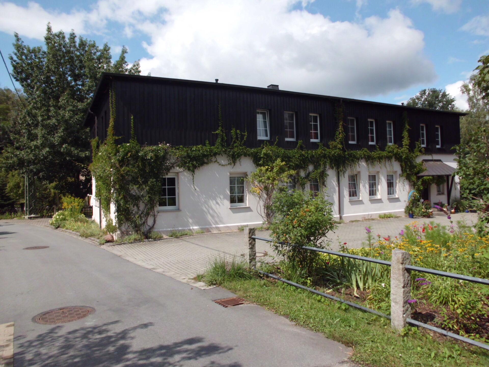 Die Pension Adriaurlaub in Crostau: Ein charmantes, zweistöckiges Gebäude mit einer weiß verputzten unteren Etage und einer dunklen Holzfassade im oberen Bereich, teilweise bewachsen mit Rankpflanzen. Davor befindet sich ein kleiner Vorgarten mit Blumen u