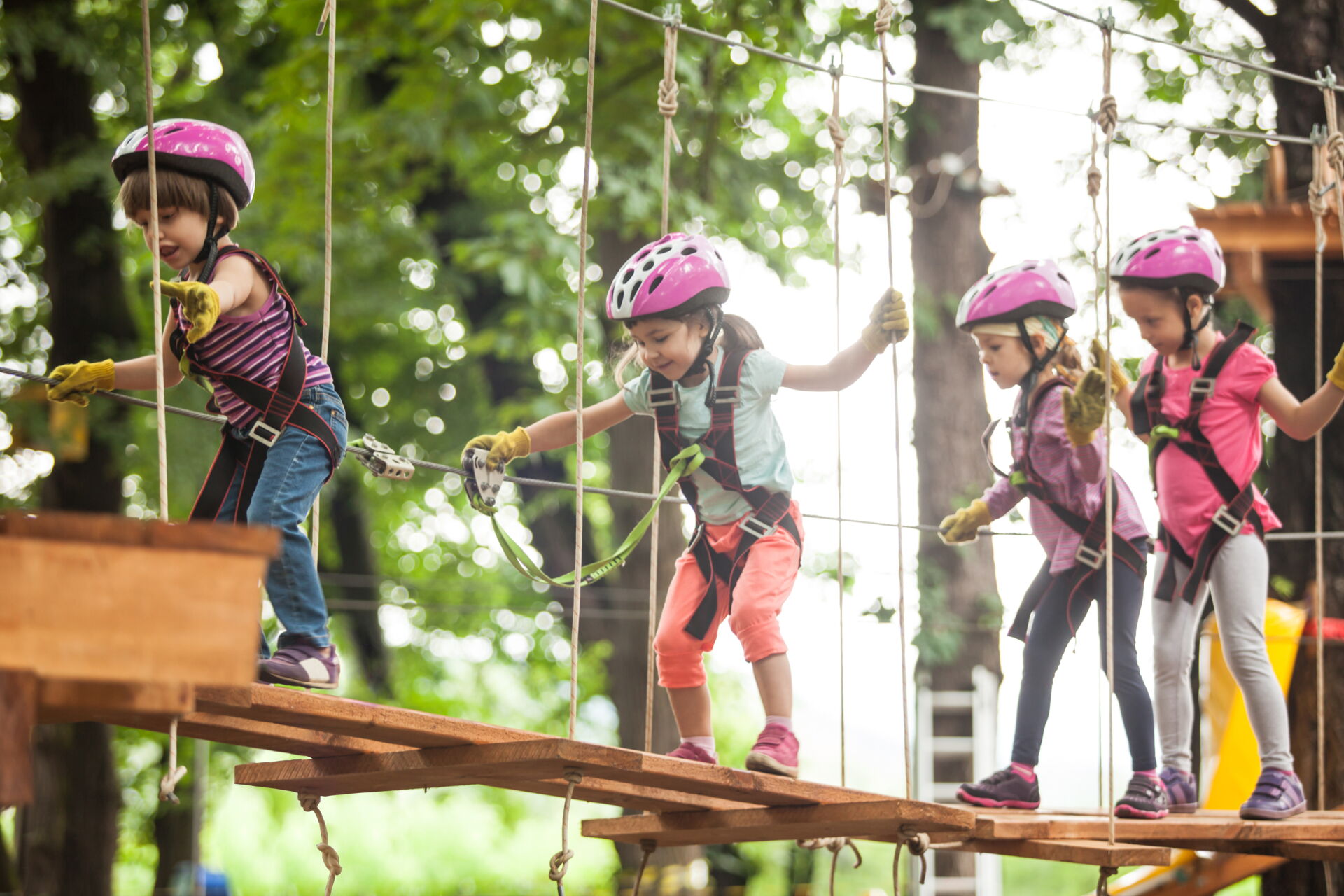 Eine Gruppe von vier Kindern mit lila Helmen und Klettergurten balanciert vorsichtig auf einem wackeligen Seilbrückenparcours in einem Hochseilgarten, umgeben von grünen Bäumen.
