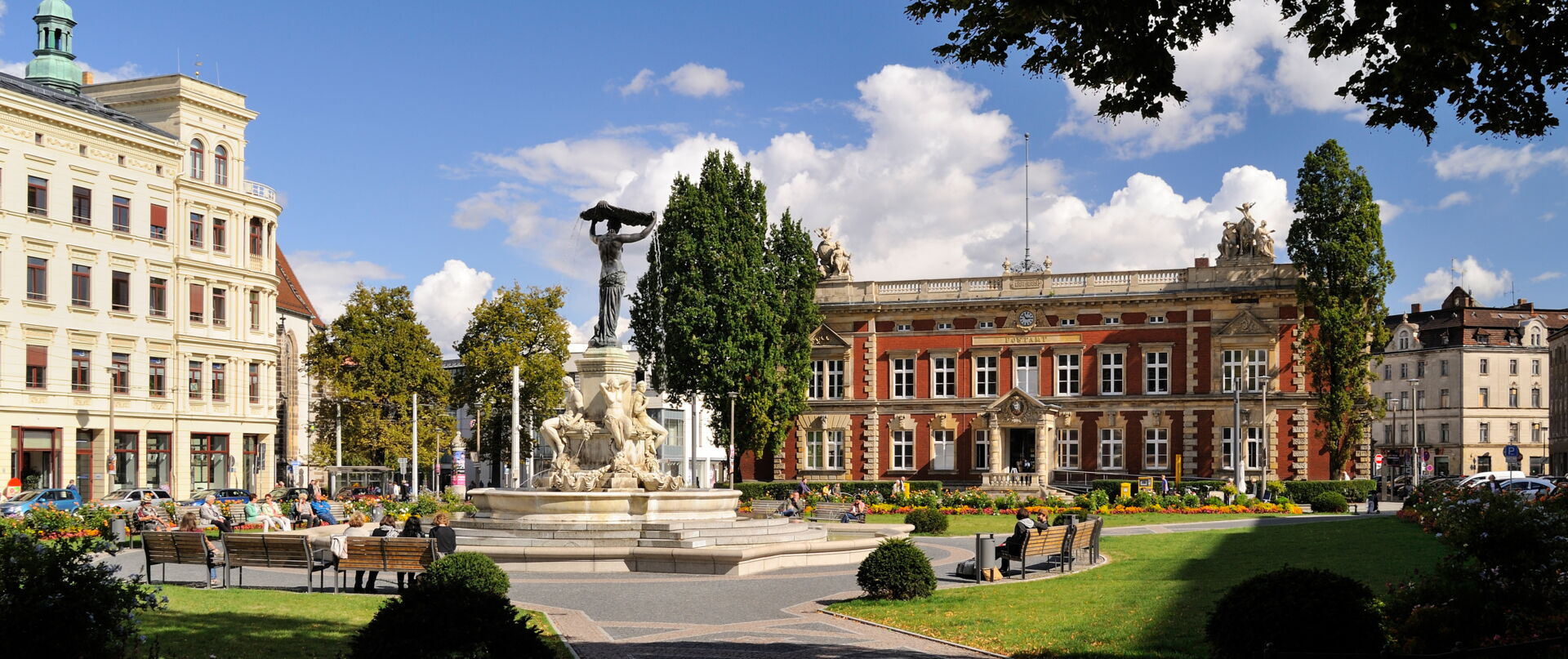 Ein sonniger Marktplatz mit einem großen Brunnen und historischen Gebäuden, Menschen sitzen auf Bänken.